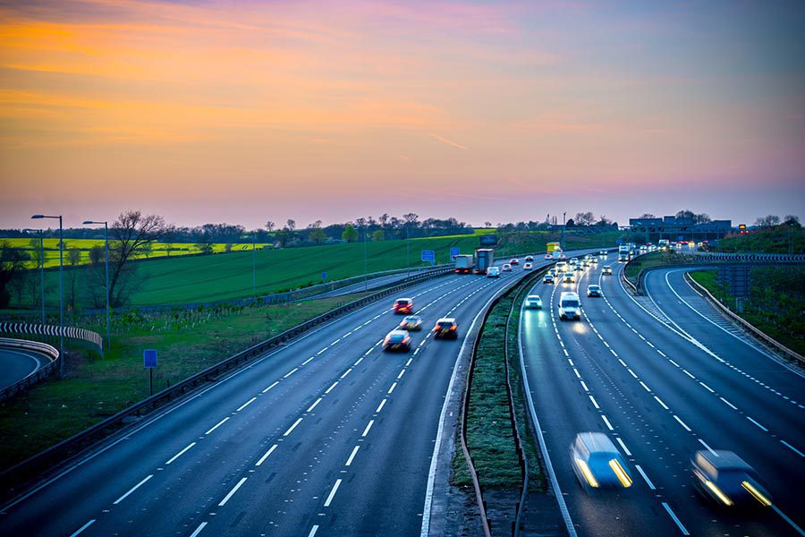 About Our Agency - Long Exposure of a Busy Highway at Sunset, Green Fields and Trees on Each Side, Cars and Trucks With Lights on Speeding Past