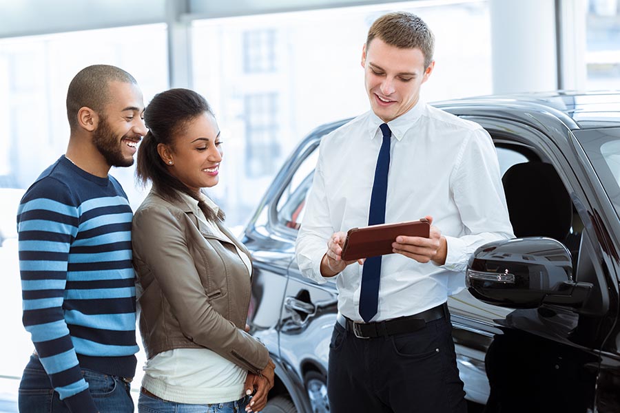 Business Insurance - Couple Shopping at Car Dealership Are Helped by a Salesman in Shirt and Tie, Showing the Couple Information on a Tablet as They Stand in Front of a Black SUV