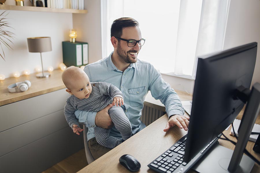 Client Center - Young Dad in His Home Office Smiles and Uses Computer With One Hand While Holding Infant Son in His Other Arm