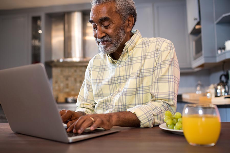 Contact Us - Senior Man Happily Using Computer at Wooden Kitchen Table With a Glass of Orange Juice and Plate of Grapes Beside Him