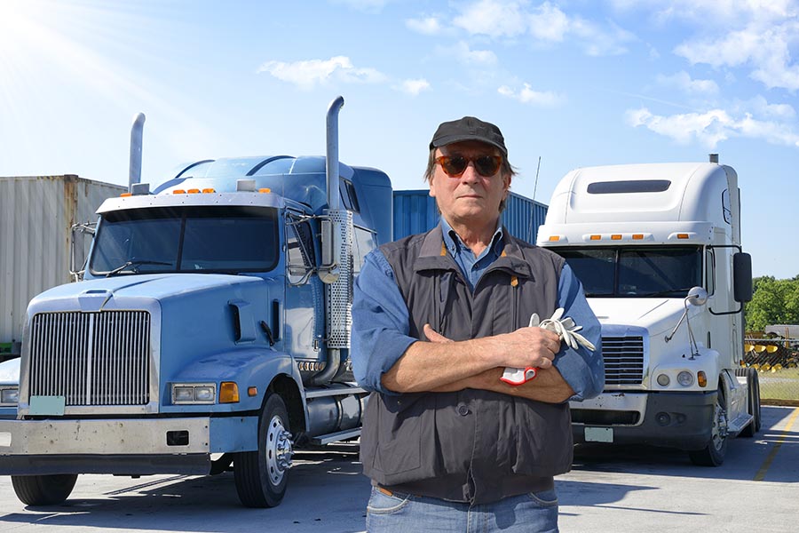 Specialized Business Insurance - Serious Truck Driver in Hat, Sunglasses, Vest and Jeans Crosses His Arms While Standing in Front of Large Trucks
